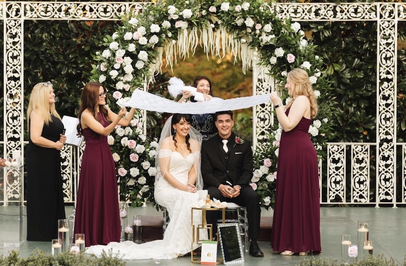 bride and groom sitting during their wedding ceremony in front of large flower covered archway at a wedding coordinated by Hearts and Souls Event Management