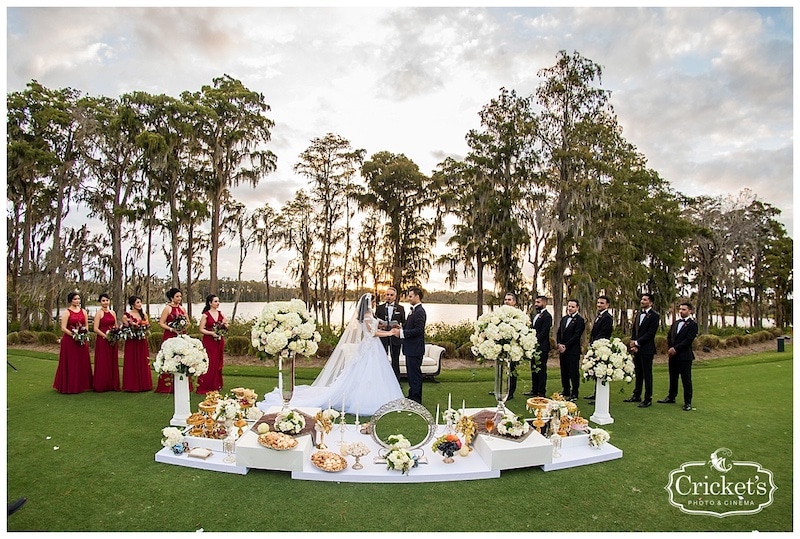 outdoor wedding ceremony on a golf course next to a lake, with a big spread of flowers and treats in front of the wedding party