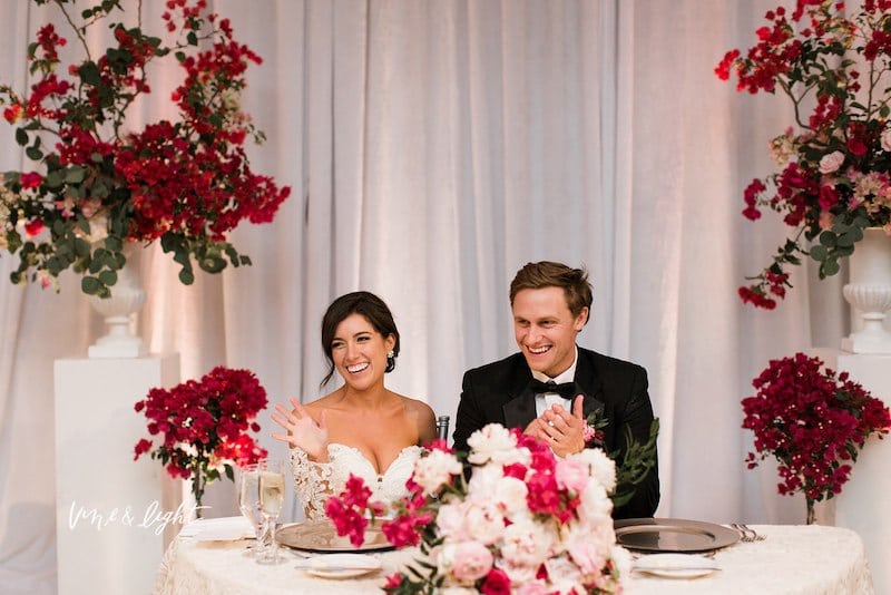 bride and groom laughing while sitting at sweetheart table surrounded by red flowers