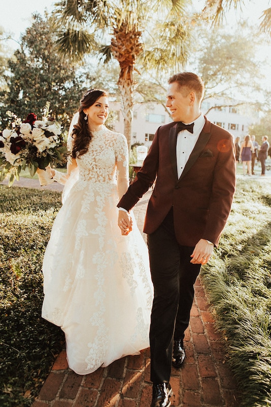bride and groom smiling at each other as they hold hands and walk down a brick path through a garden