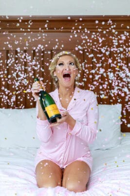bride in a pink robe kneeling on the bed as confetti falls around her and she holds a bottle of champagne