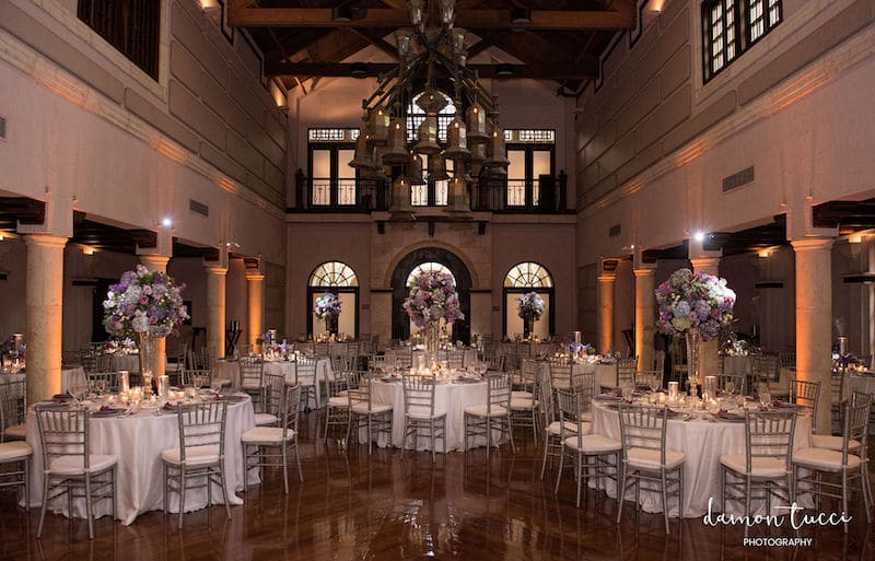 large room set up for wedding reception with tall flower centerpieces and uplights on all of the columns