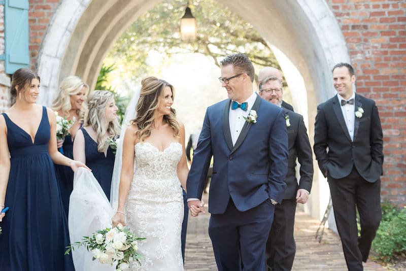 bride and groom holding hands and smiling at each other as they walk on a brick path with their bridal party