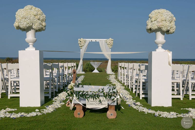 outdoor wedding ceremony overlooking the ocean with a wagon that has a sign that reads Daddy here comes Mommy.