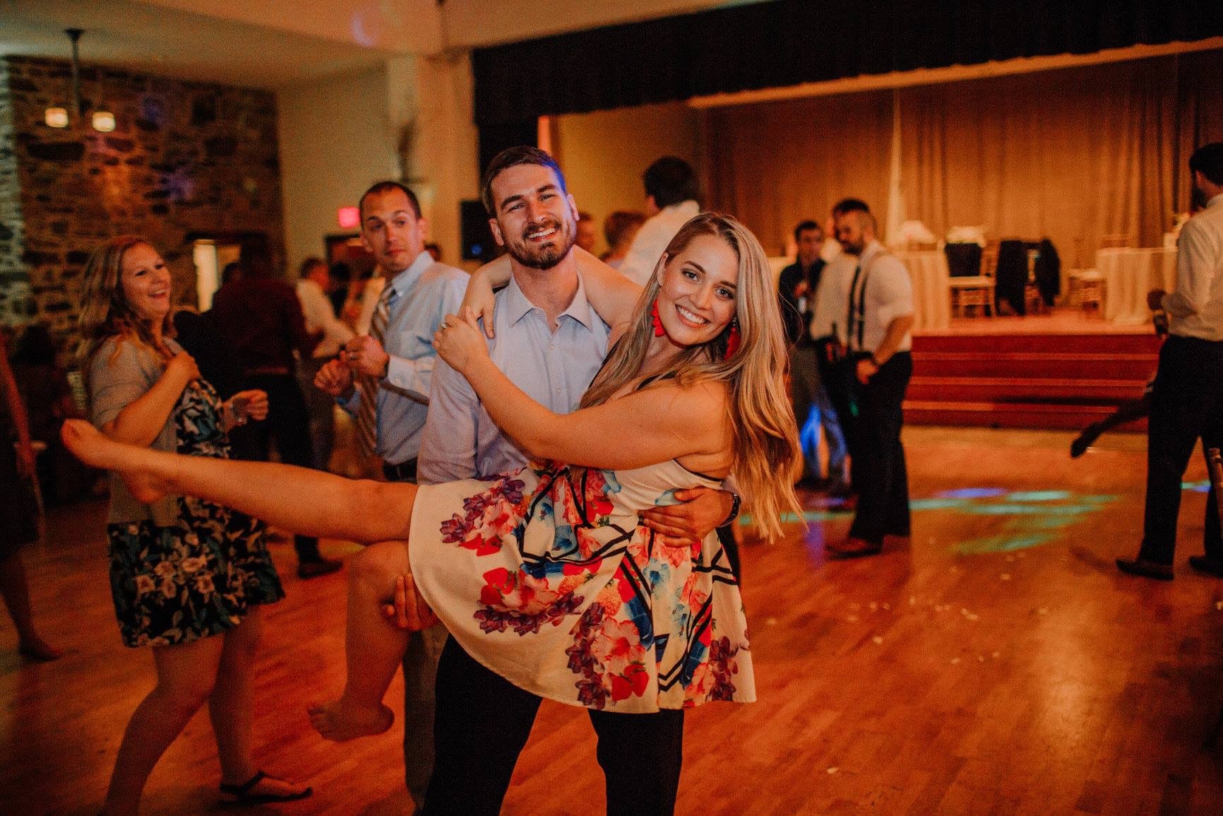 woman wearing a dress and woman a button down shirt and pants pose for a picture as the man picks her up on the dance floor at an event