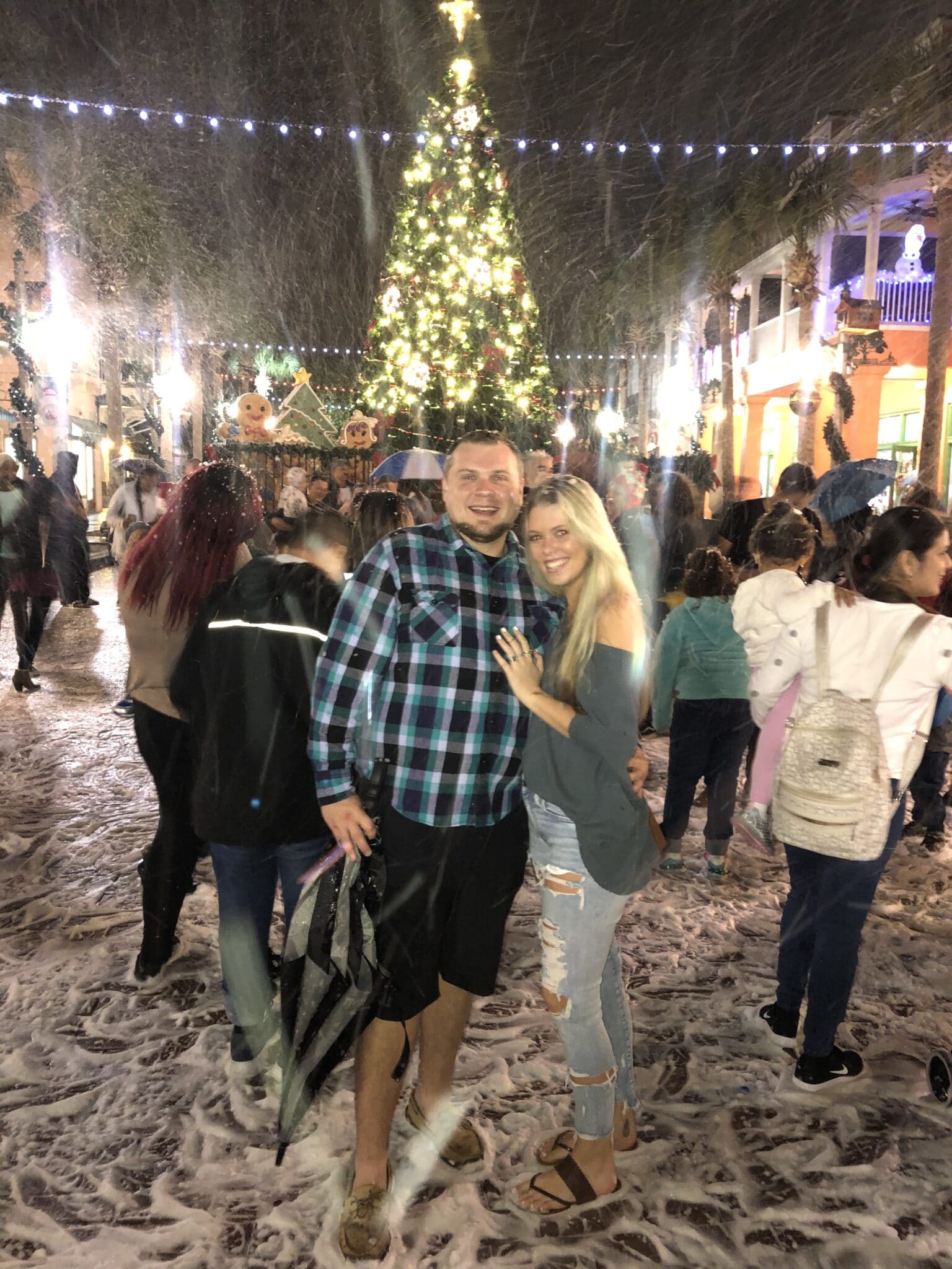 couple standing together in front of a large christmas tree with people surrounding them