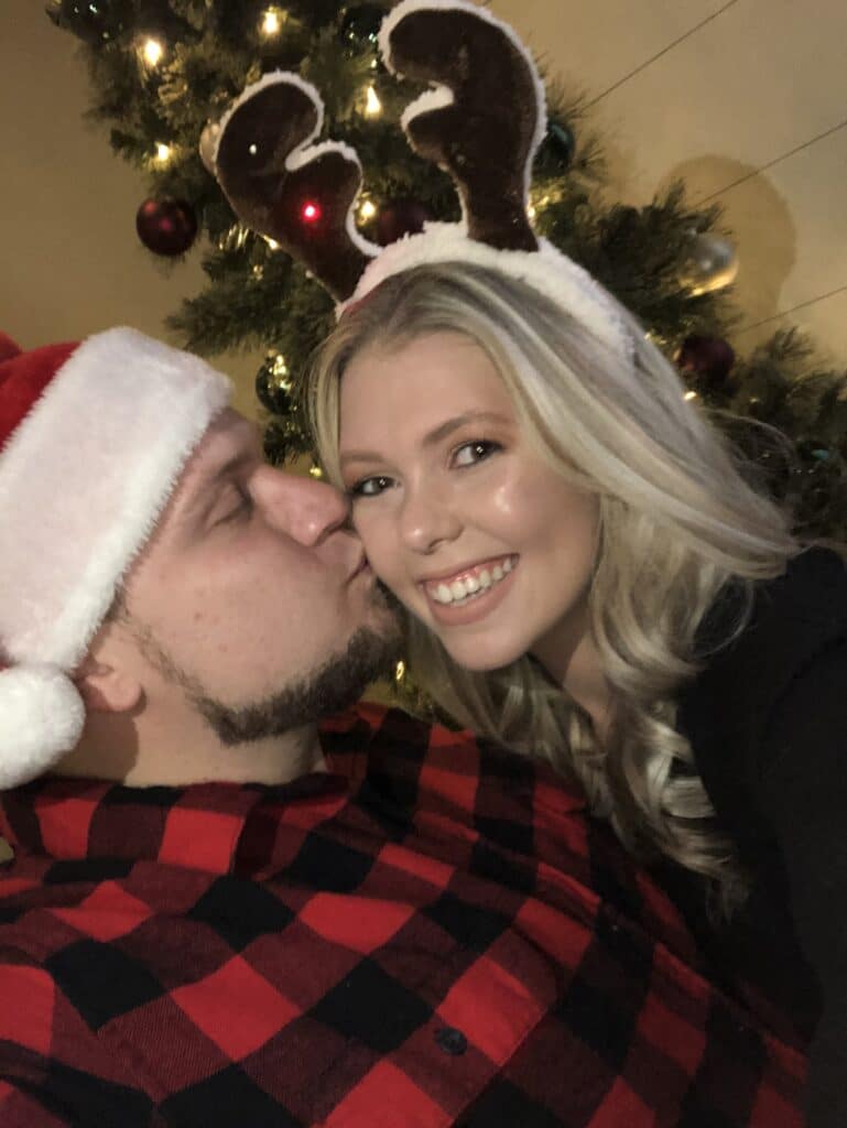 groom to be with a santa hat on kissing the bride to be on the cheek while she smiles