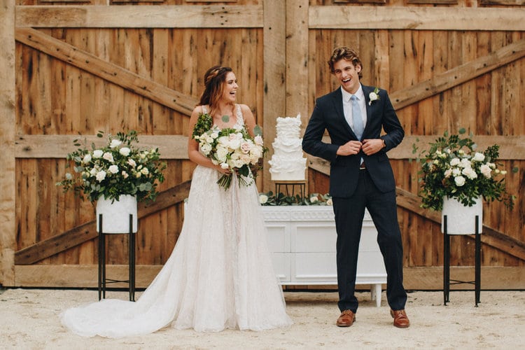 bride and groom laughing in front of large barn doors with white flowers