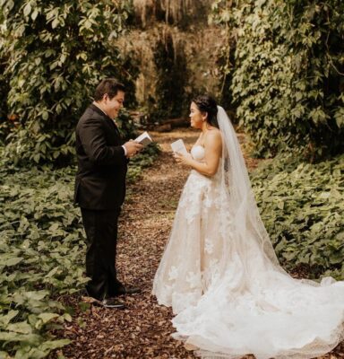 bride and groom smiling at each other while they read notes to one another on trail in woods