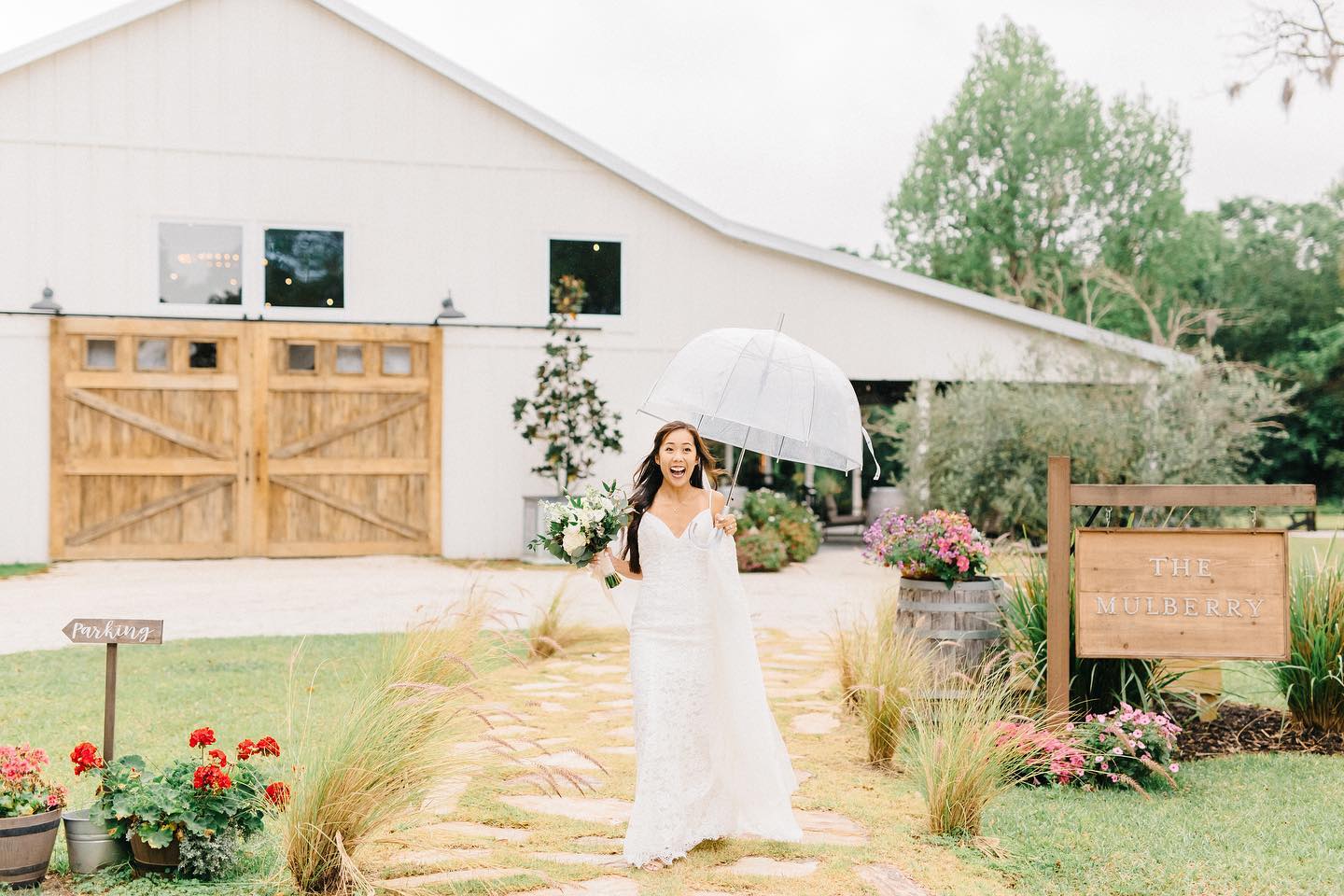 bride holding bouquet and umbrella while walking down path in front of white barn