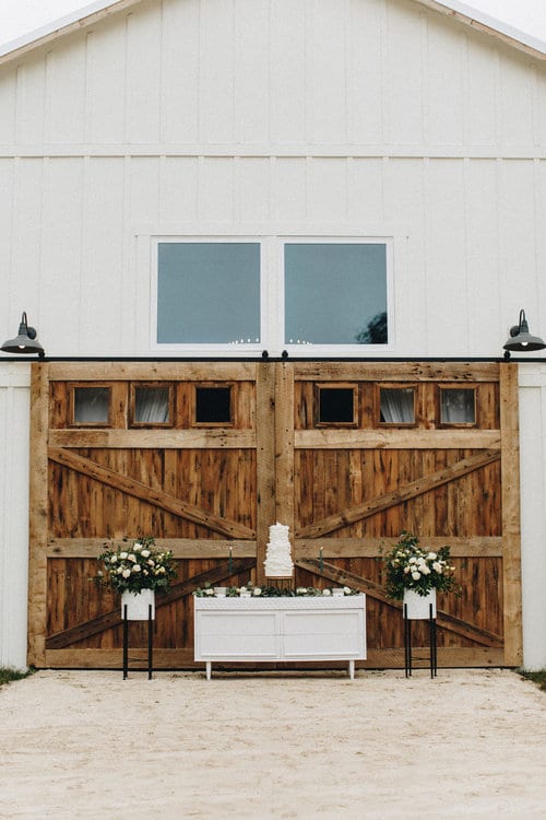 white barn with large wooden barn doors, with table and wedding cake in front of it