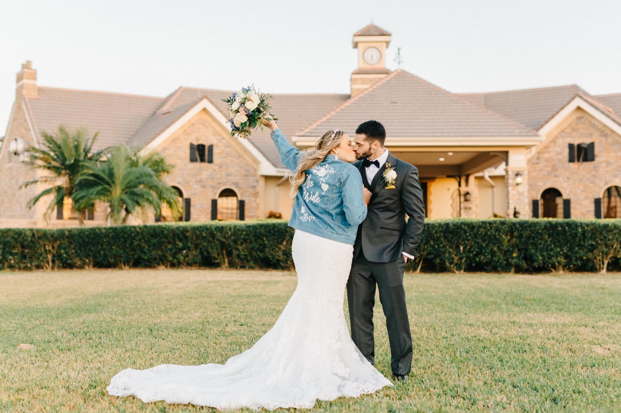 bride wearing a custom jean jacket kissing her new husband and holding her flower bouquet in the air at Eagle Creek Golf Club