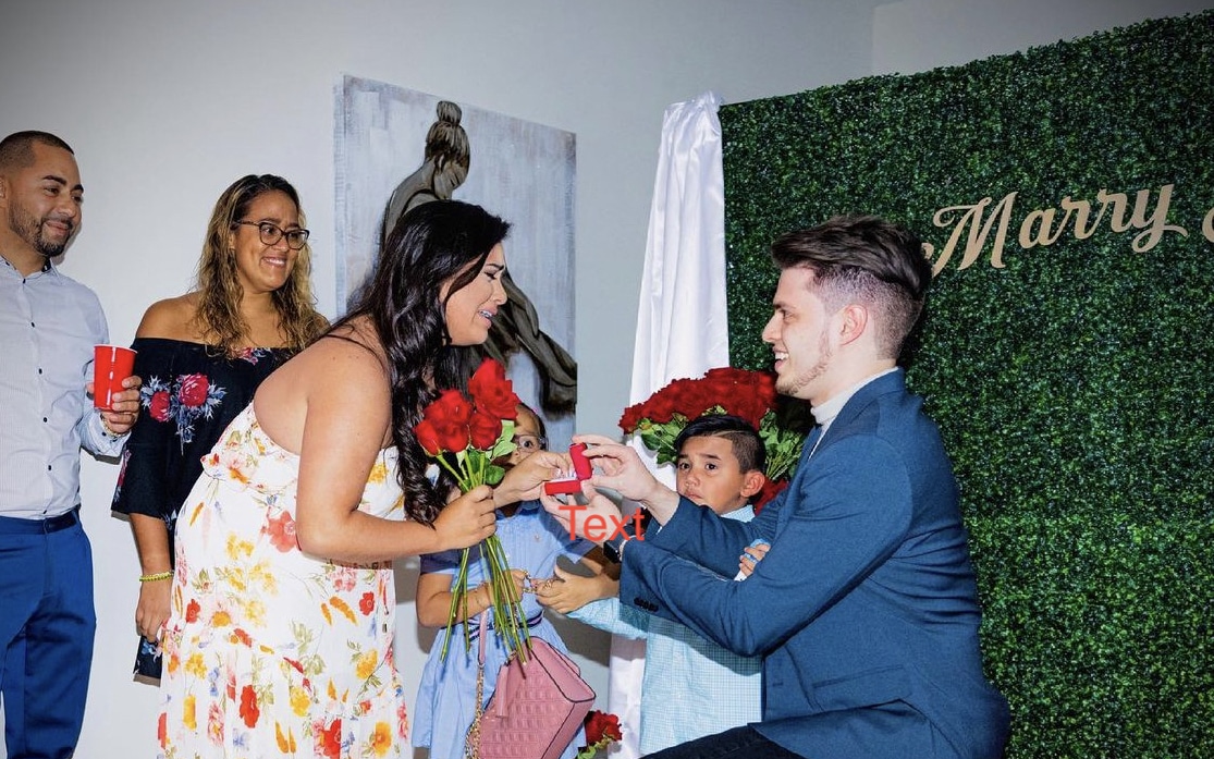 groom to be on one knee proposing to the bride to be with friends and family all around