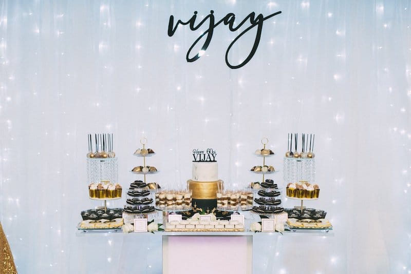 large dessert table filled with cakes, cookies, and other desserts set up in front of a white curtain with string lights