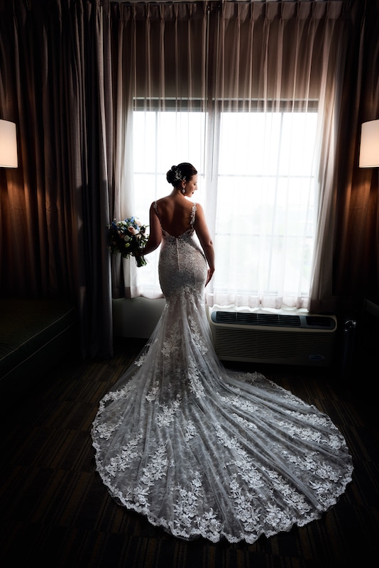 bride standing in her wedding dress in front of open window by Lazzat Photography