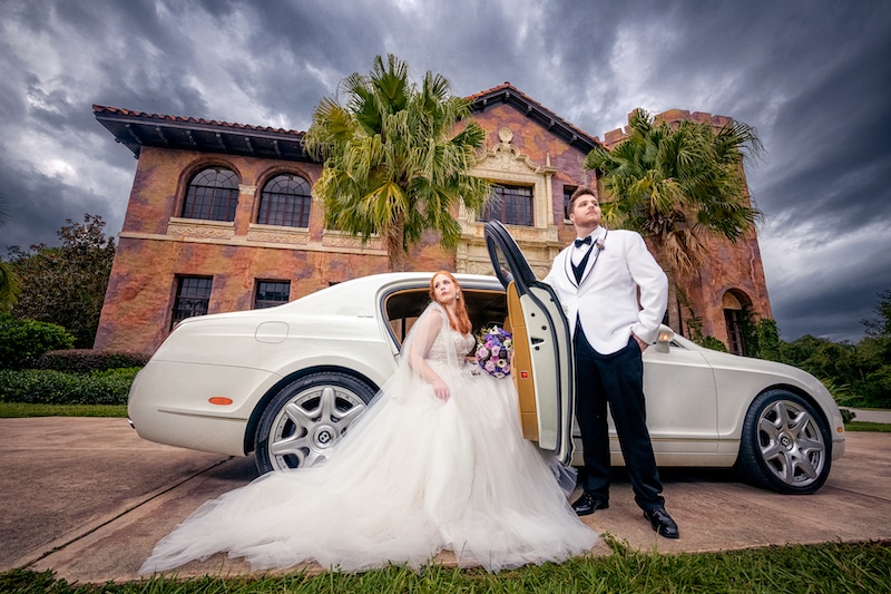 groom holding the door while his bride gets out of a white car in front of a large mansion by Lazzat Photography