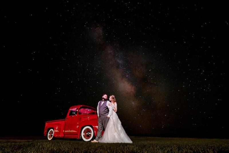 bride and groom standing in a field outside at night next to a red truck with the milky way galaxy visible in the night sky by Lazzat Photography