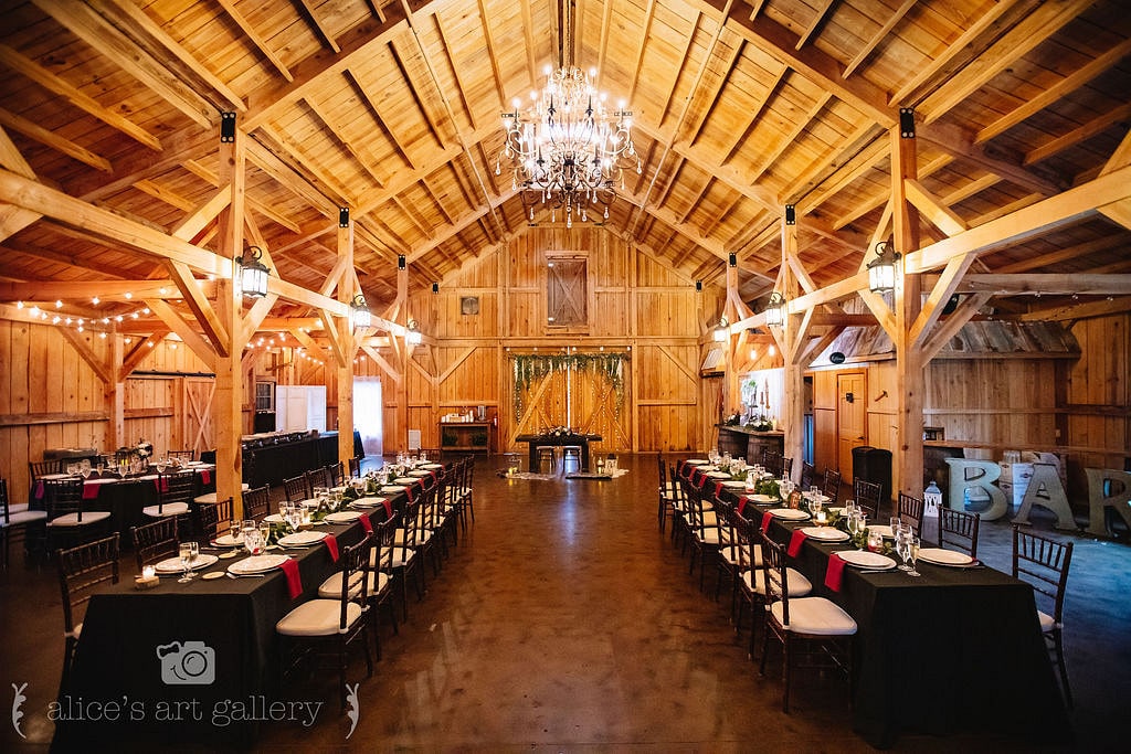 wedding reception space inside barn with giant chandelier handing with long rectangle tables on both sides and a sweetheart table at the front in front of two barn doors