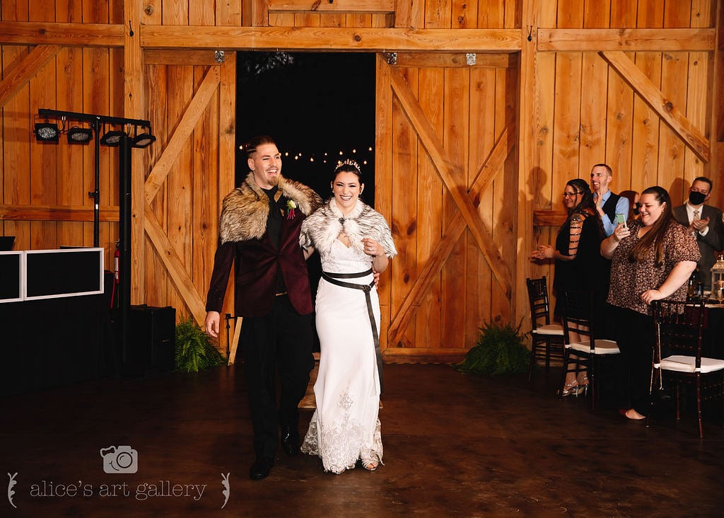 couple on wedding day walk into wedding reception holding hands and smiling through barn doors with dj booth to the left and guests on the right