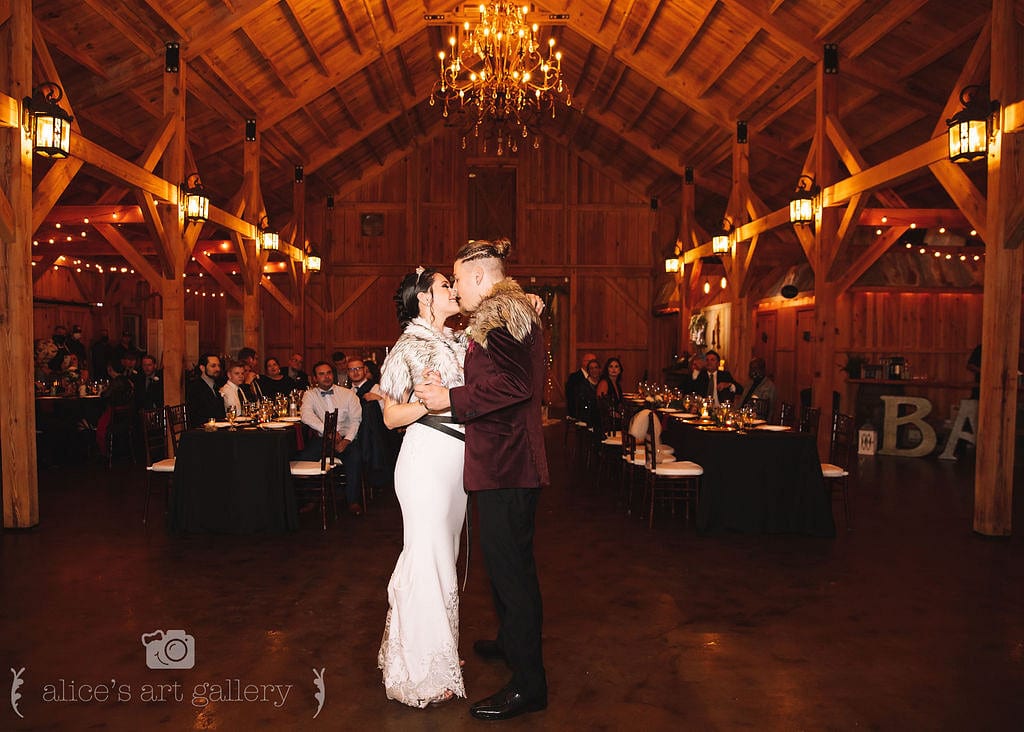 bride and groom stand together holding hands and touching faces in dimly lit barn dancing as guests watch