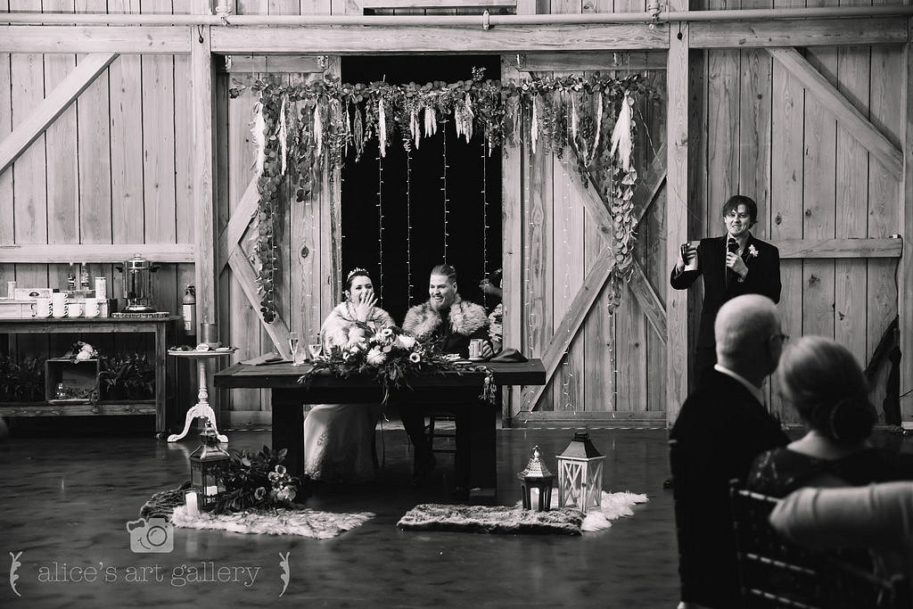 black and white image of bride and groom sitting at sweetheart table at wedding reception while groomsmen stands to the right holding microphone and speaking