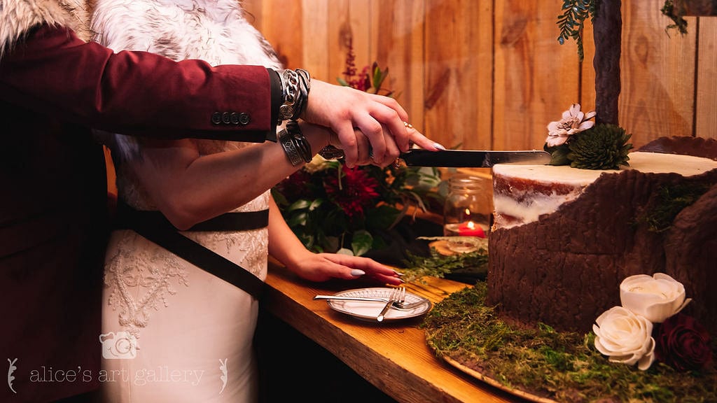 man holds hand of woman holding large knife about to cut into cake decorated with chocolate wood bark and cake plate with forks sits in front of it