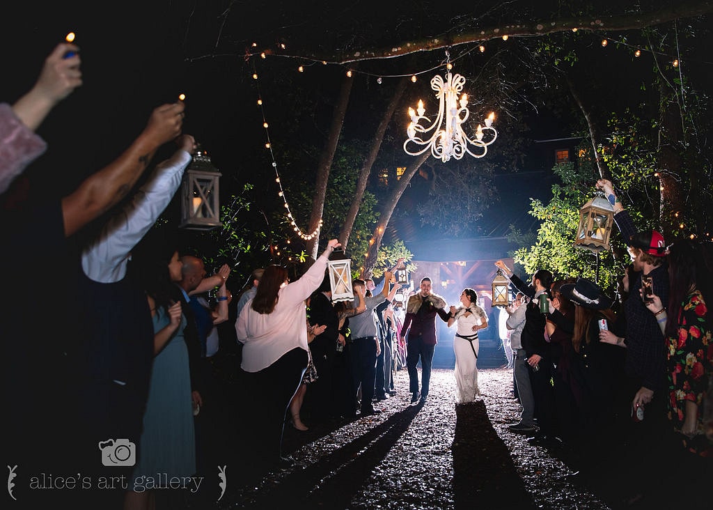 couple on wedding day walk down path outside at night while wedding guests hold out lanterns into the path and chandelier hangs above them