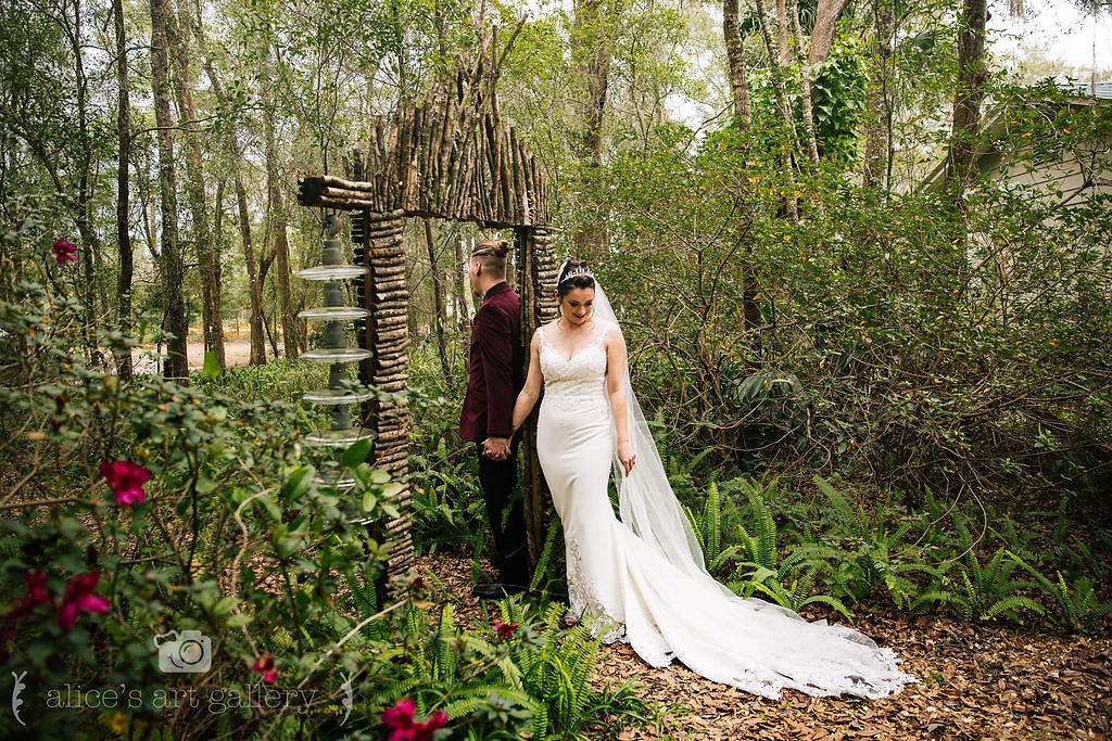 bride and groom hold hands while facing away from each other outside in garden of forrest and wooden decoration structure