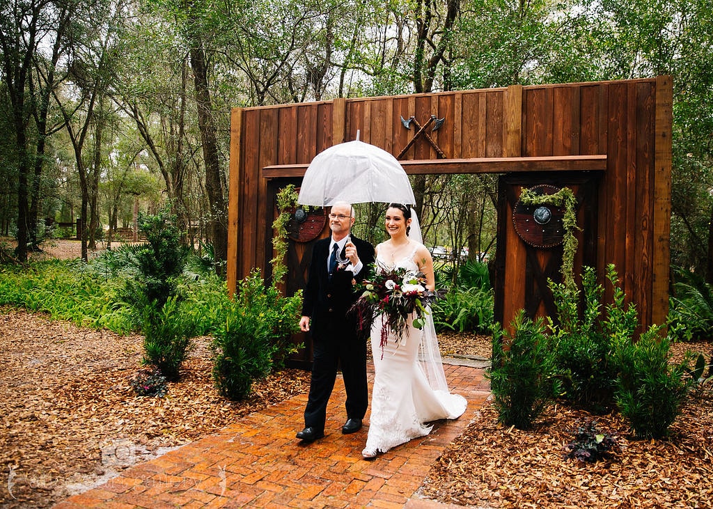 bride holds bouquet and walks with father down the aisle while holding clear umbrella on wedding day in front of large wooden barn doors