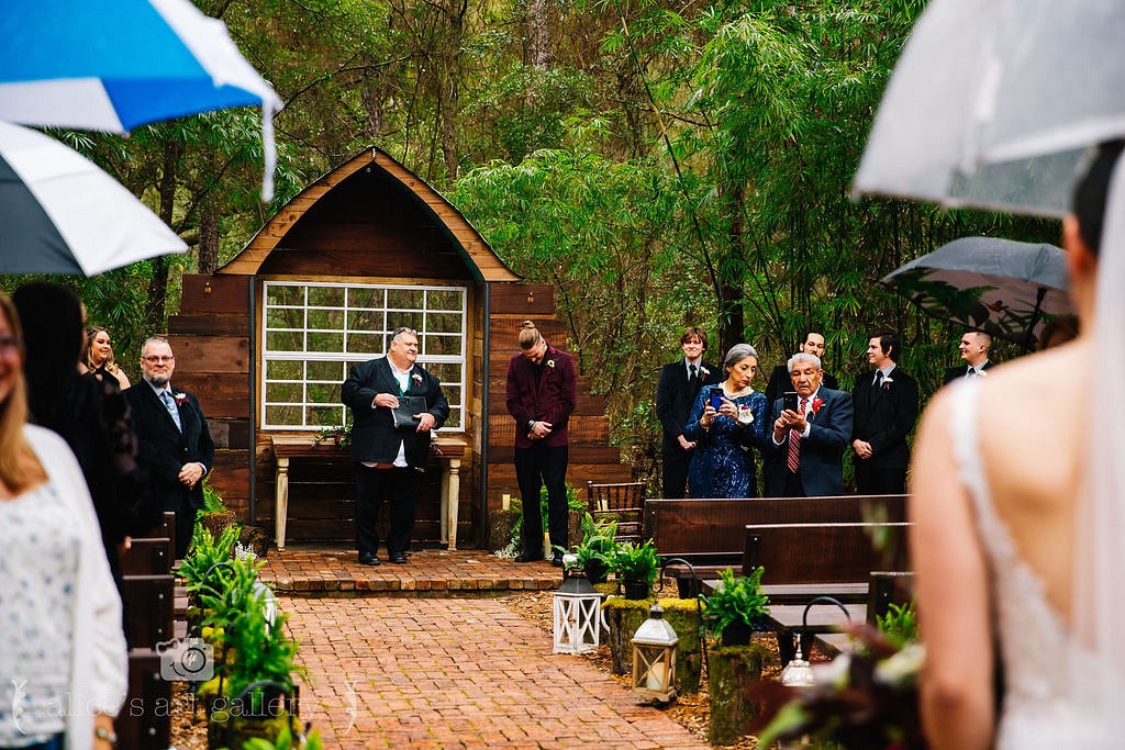 groom stands at altar with officiant while guests turn to watch the bride walks down the aisle while holding umbrellas