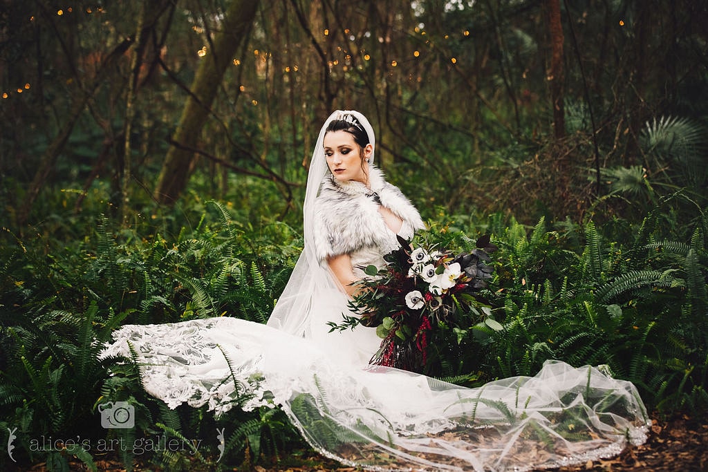 bride sits in greenery outside with wedding dress and veil spread out around her and holding her bouquet in front of her as she looks off to the left