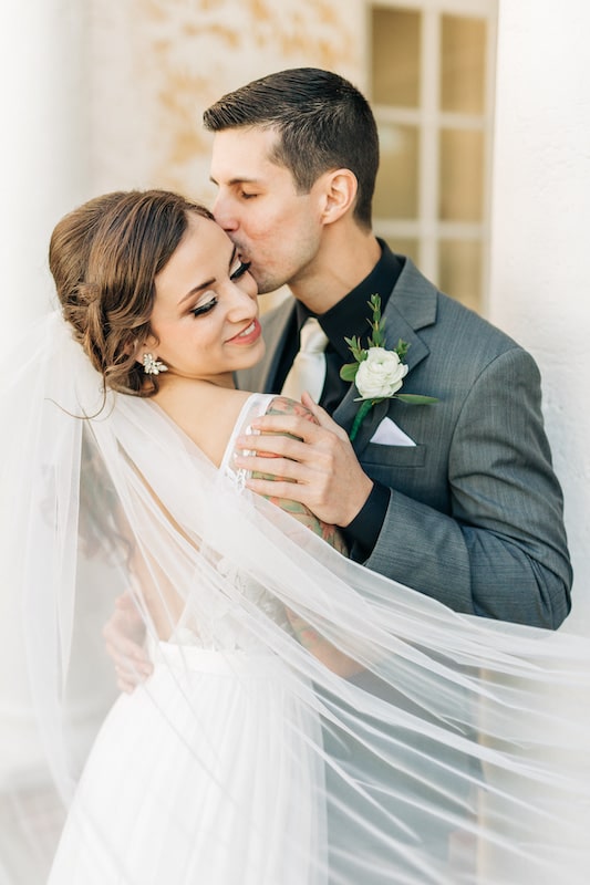 groom kissing his bride on her cheek while both are wrapped in her veil with planning by Weddings by Kasey