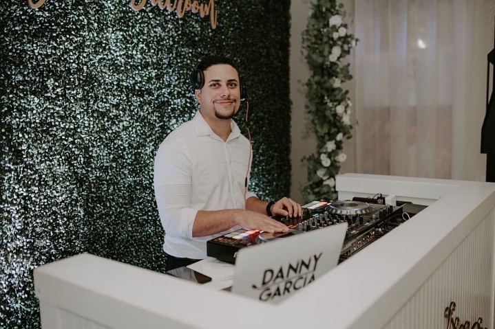 wedding dj with headphones on smiling behind dj booth at wedding with green hedge wall behind him