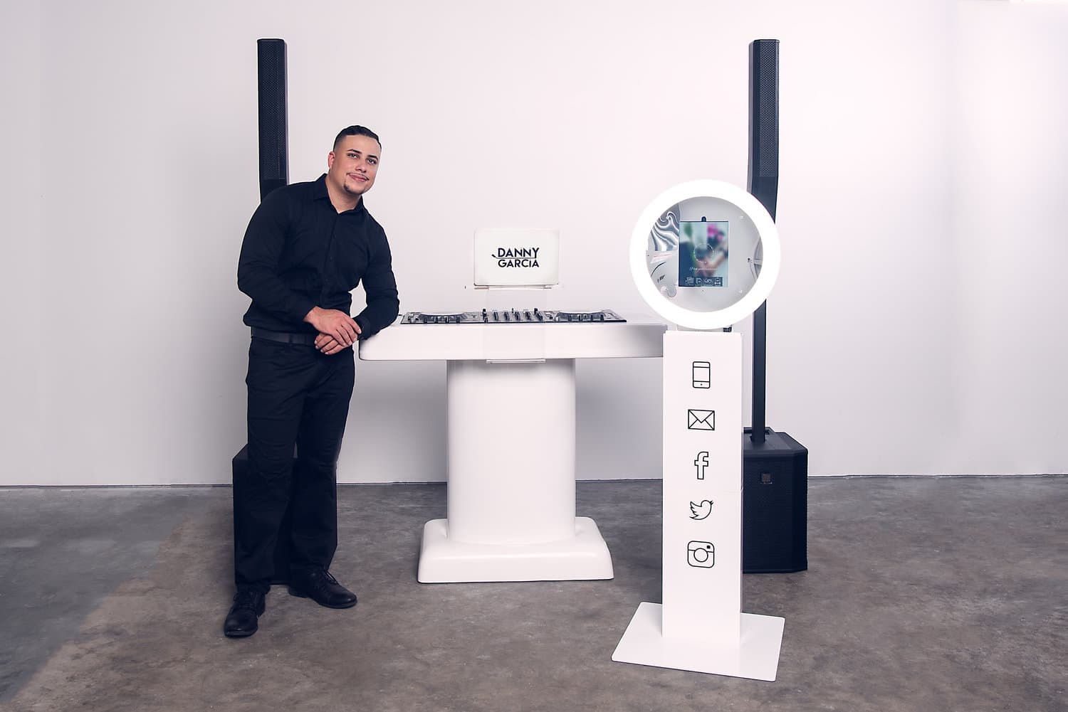 man stands dressed in all black leaning against white dj booth with photo booth stand in front of it and two speakers on each side of booth