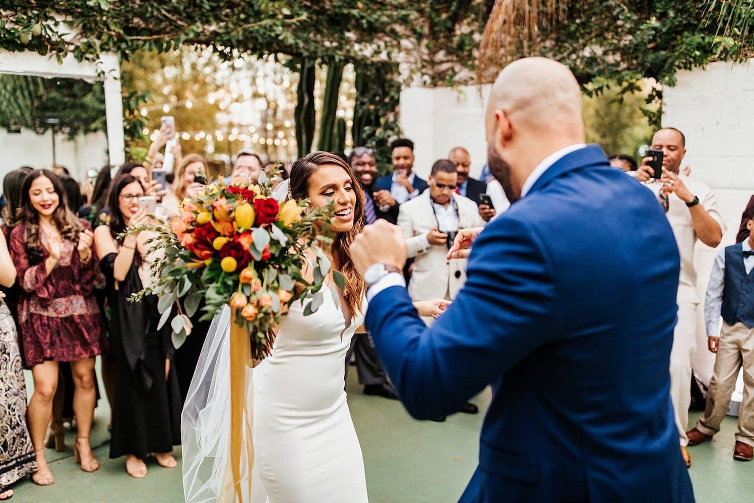 bride holding bright colored bouquet of flowers dancing next to groom with guests standing around them