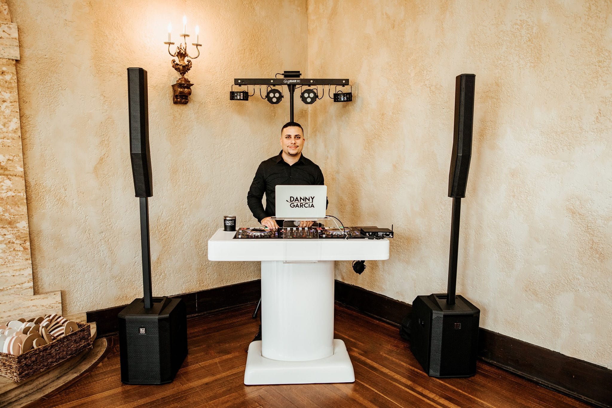 dj stands behind simple white dj booth stand with speakers on both sides and lights on a stand behind him in the corner of a room