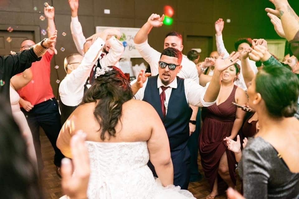 bride and groom dance in center of friends and family on wedding day while groom wears sunglasses