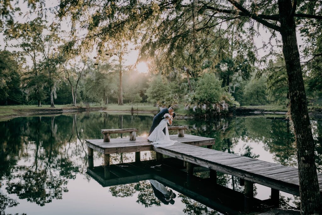 groom dipping and kissing his bride while standing on a small dock near a pond