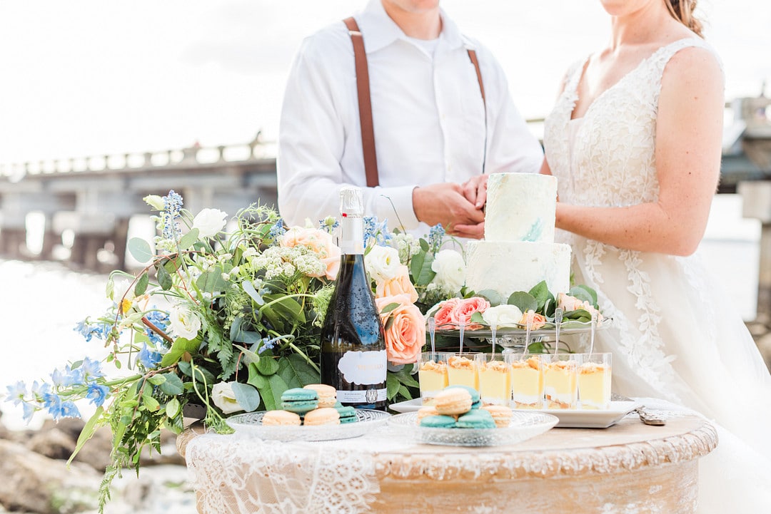 the dessert table with macrons, pudding, champagne and florals and the couple standing in the background