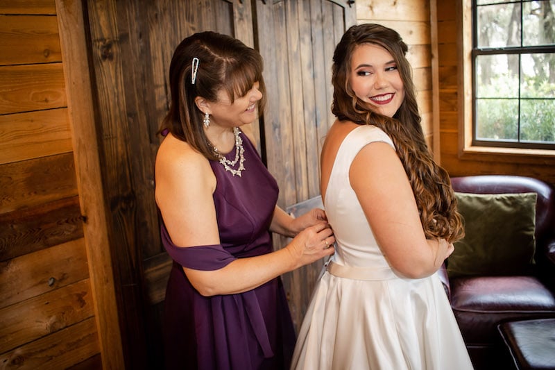 bride smiling and looking back at her mother who is helping her get into her wedding dress, SMO Photography