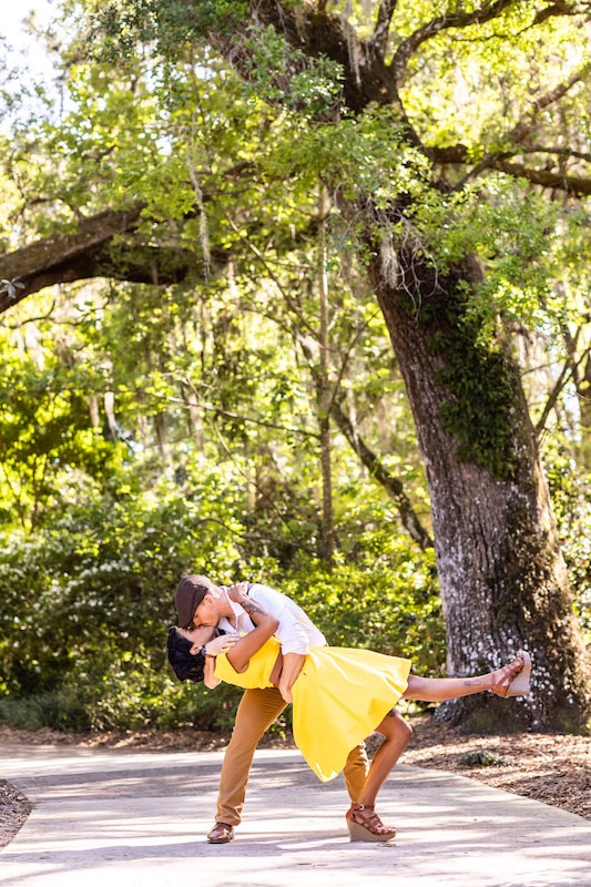 man dipping and kissing a woman in a yellow dress while standing on a path under a large tree, SMO Photography