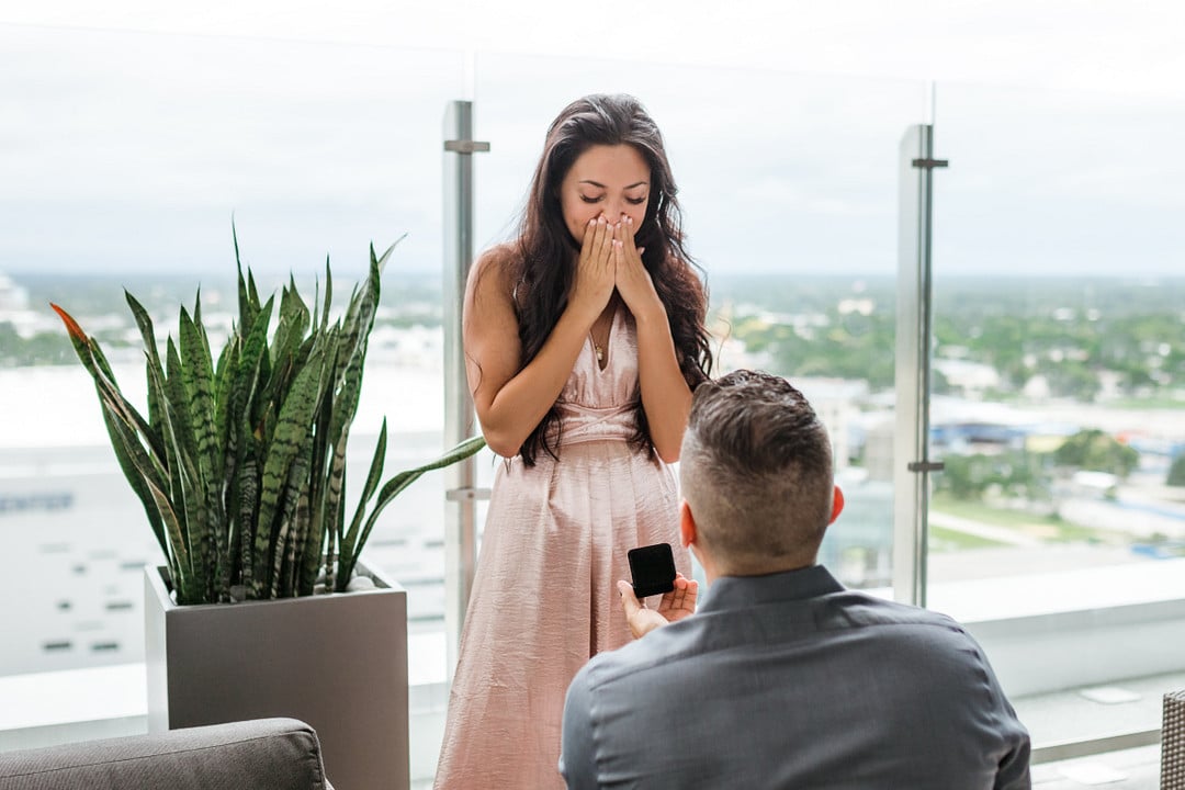 groom to be on one knee proposing to his girlfriend with her hands covering her mouth in shock