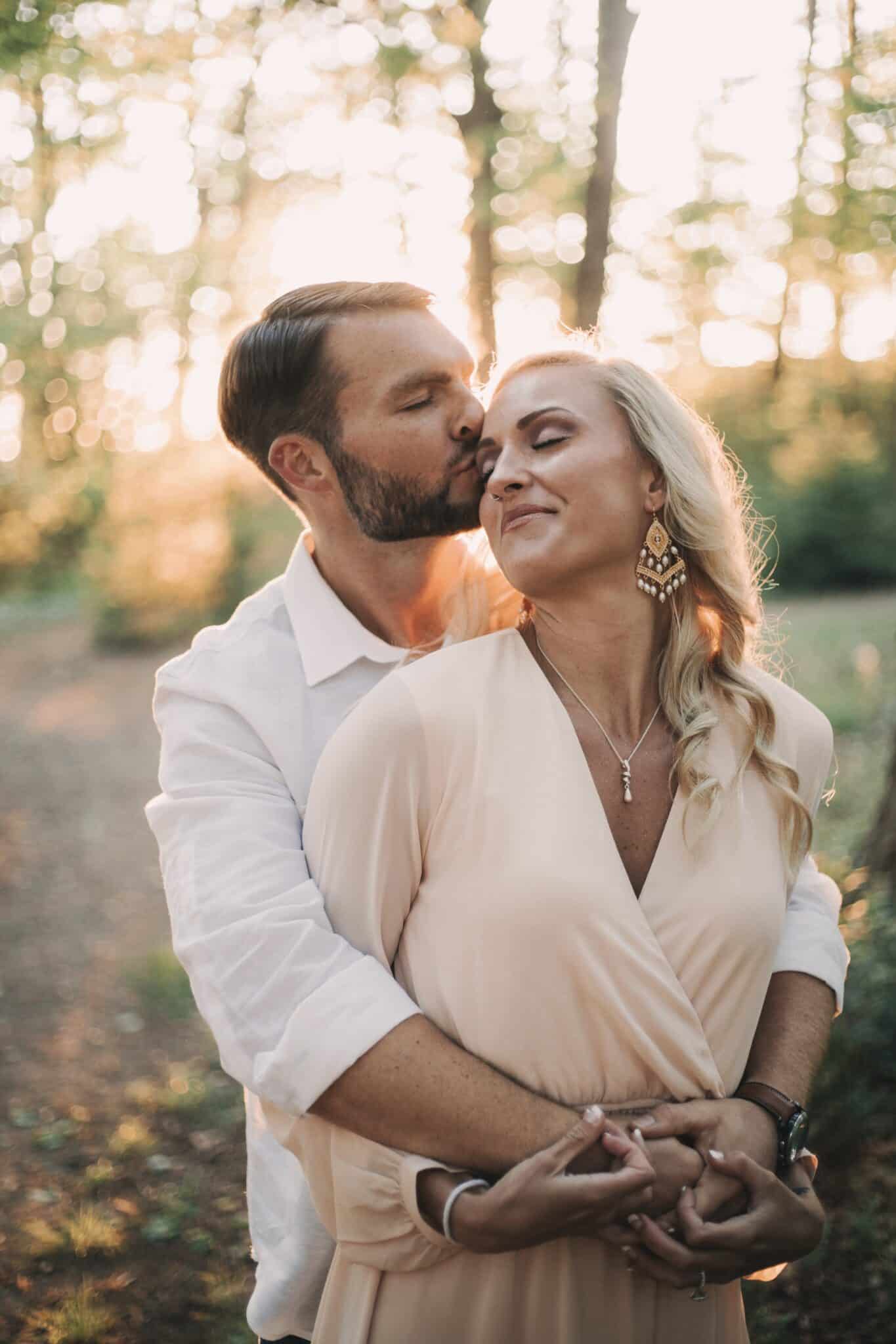 groom to be kissing his bride to be's forhead in the woods after their staycation marriage proposal