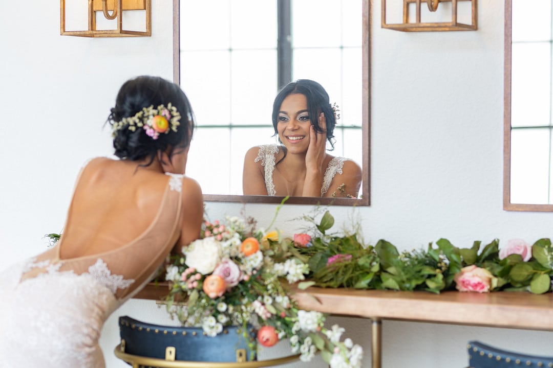 bride at the summer bright wedding inspiration shoot looking in the mirror and smiling with flowers on the table and in her hair
