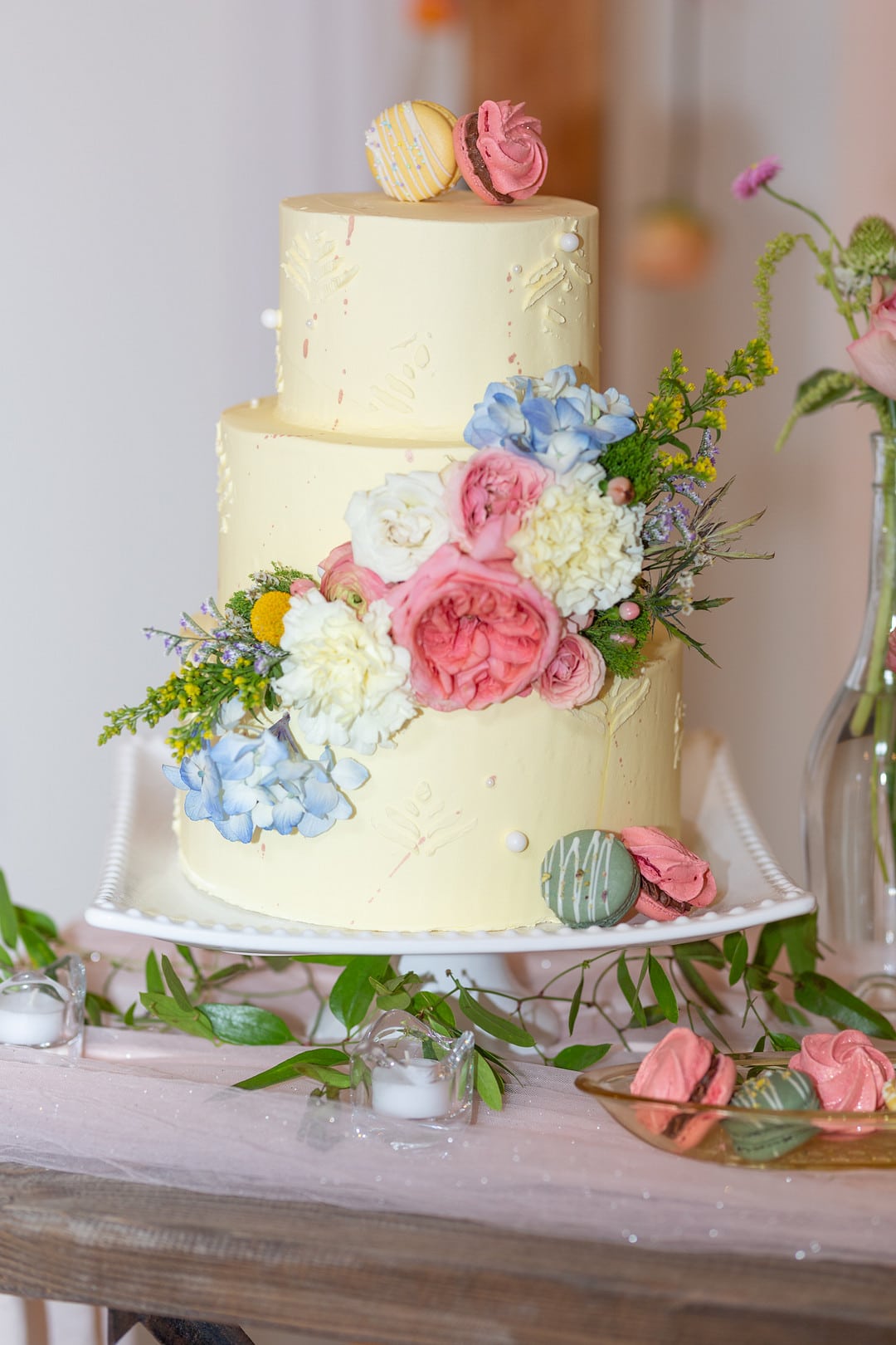 a three-tiered cake with pink, blue and white flowers surrounded by macarons and florals