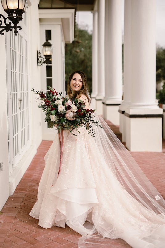 bride standing outside of large building with huge white pillars while smiling and holding her flower bouquet