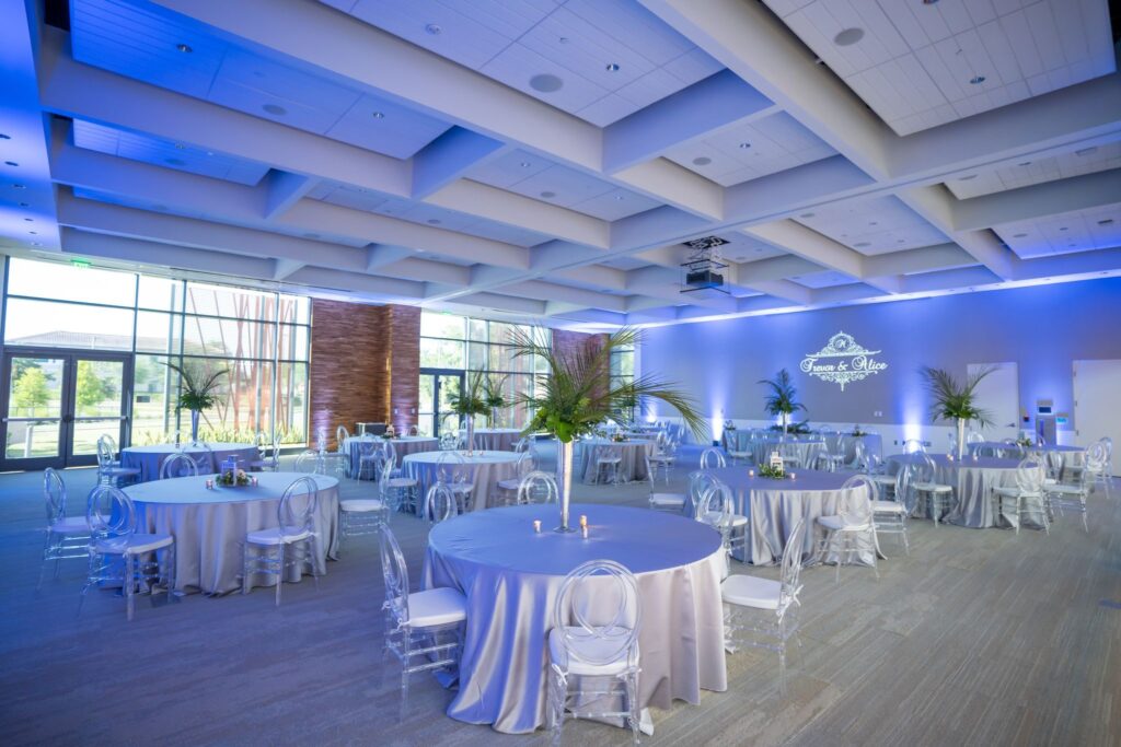 large room at The Wellvue set up for wedding reception with several tables, blue uplights, and a personalized gobo projector