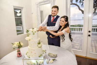 bride and groom cutting into cake with white flowers on each tier in ballroom facing lake