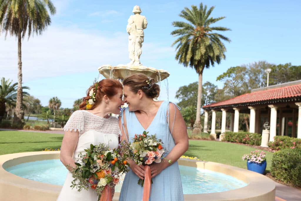 bride and bridesmaid smiling at each other while standing in front of fountain