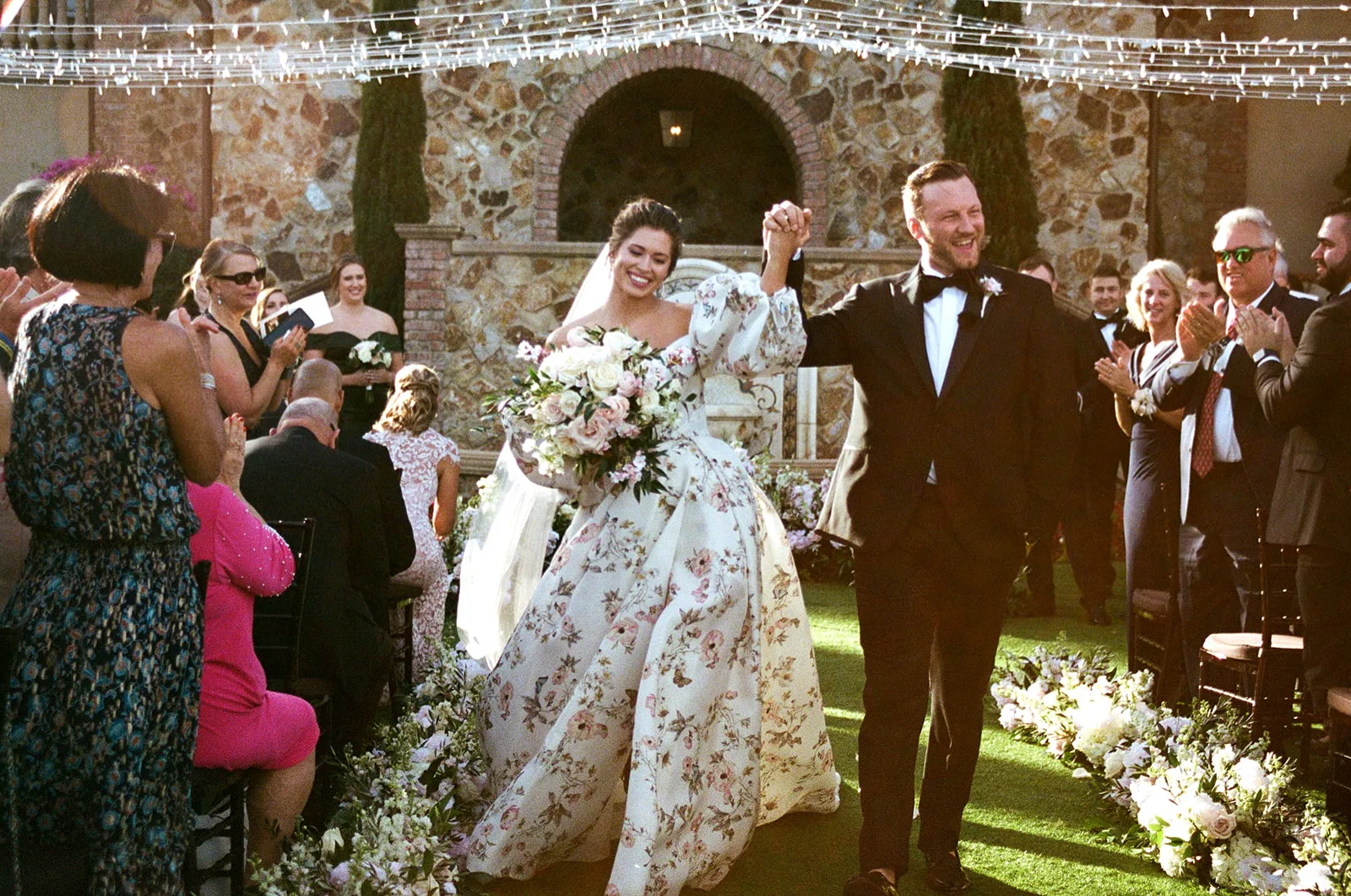 Bride in a floral wedding gown and groom in a classic tuxedo walk down the aisle smiling and hand-in-hand beneath twinkling lights, at a romantic outdoor ceremony at Bella Collina planned by Anna Christine Events.